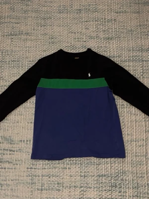 Långärmad topp från Polo Ralph Lauren - Snygg långärmad topp från Polo Ralph Lauren med svart överdel, grön rand och marinblå nederdel. Klassisk logga broderad på bröstet. Tillverkad i mjuk bomull, perfekt för en chill och stilren look.