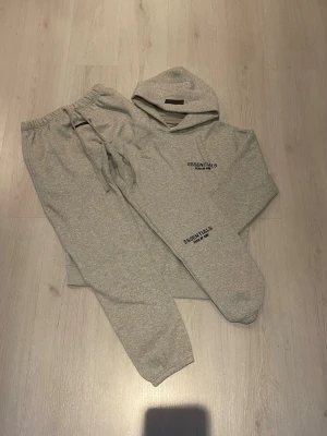 Grått loungewear-set Essentials - Snabb leverans. Pris kan vi prata om. Det är en Mysigt grått loungewear-set från Fear of God Essentials med hoodie och matchande byxor. Setet har svart logotyptryck på både tröjan och byxan. Tillverkat i mjukt bomullsmaterial med dragsko i midjan och huva för extra komfort.