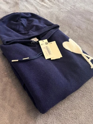 Ami Paris Hoodie  - Snygg mörkblå hoodie från Ami Paris med huva och dragsko. På bröstet finns ett vitt hjärta med bokstaven A, vilket är typiskt för märket. Tillverkad i mjukt material som känns skönt mot huden. Perfekt för en avslappnad och trendig stil.