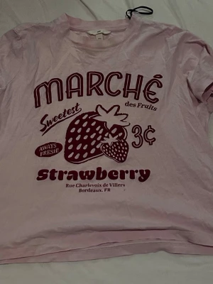 Ljusrosa t-shirt med jordgubbstryck - Söt ljusrosa t-shirt med rött tryck framtill som föreställer en jordgubbe och texten 'Marché des Fruits' och 'strawberry'. T-shirten har rund hals och korta ärmar. Perfekt för dig som gillar gulliga och lekfulla plagg.