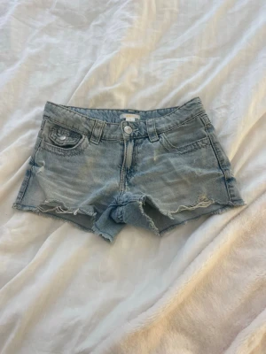 Ljusblå jeansshorts med slitningar - Säljer ett par ljusblå jeansshorts från Ginatricot med råa kanter och slitna detaljer. Shortsen har broderade bakfickor med knappar och klassisk femficksdesign. Perfekta för varma dagar och en avslappnad look.