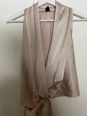 Champagnefärgad omlottopp i satin - Säljer en champagnefärgad omlottopp i satin med knyt i midjan. Toppen är ärmlös och har en djup v-ringning. Perfekt för dig som gillar en elegant och trendig look.