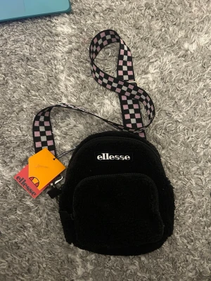Svart axelväska från Ellesse - Säljer en svart axelväska från Ellesse i mjukt teddy-material. Väskan har ett schackrutigt axelband i svart och rosa, en framficka med dragkedja och Ellesse-logga framtill. Perfekt storlek för det viktigaste och riktigt snygg detalj till din outfit.