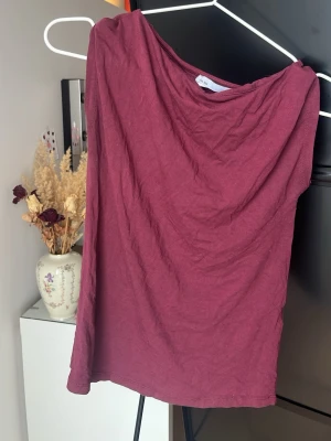 Vinröd oneshoulder topp - Säljer en vinröd oneshoulder topp i mjuk bomull. Toppen är ärmlös på ena sidan och har en snygg asymmetrisk skärning som ger en trendig look. Perfekt att styla med jeans eller kjol för en cool vibe.