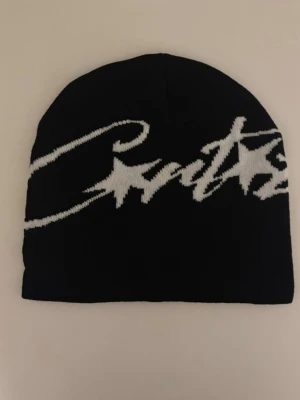 Svart mössa med vit logga - Snygg svart mössa med stor vit logga i grafisk stil över hela framsidan. Tillverkad i mjukt stickat material som sitter skönt på huvudet. Perfekt accessoar för en streetwear-look.