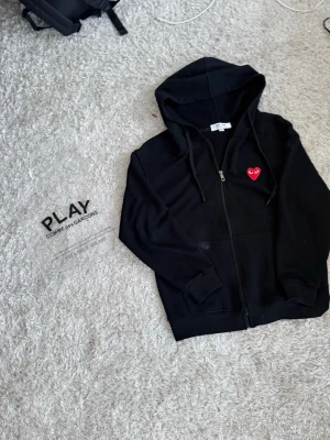 Svart hoodie Comme des Garçons Play xs - Svart hoodie med dragkedja från Comme des Garçons Play. Klassisk design med huva och fickor, samt det ikoniska röda hjärtat med ögon broderat på bröstet. Perfekt för en clean och stilren streetwear-look.