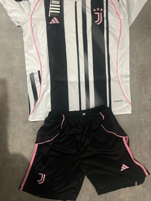 Juventus matchställ från Adidas - Juventus matchställ med tröja och shorts från Adidas. Tröjan är vit med svarta ränder och rosa detaljer, samt Juventus-logga och tre stjärnor på bröstet. Shortsen är svarta med rosa detaljer och Juventus-logga. Tillverkat i lätt och ventilerande material.