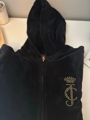 Svart velour hoodie Juicy Couture - Säljer en svart velour hoodie från Juicy Couture Black Label med dragkedja och guldfärgade detaljer. På bröstet finns ett JC-emblem med krona i guldnitar. Hoodien har en mjuk känsla och klassisk huva. Perfekt för en chill och lyxig look. Tröjan är några säsonger gamla, alltså vintage. 