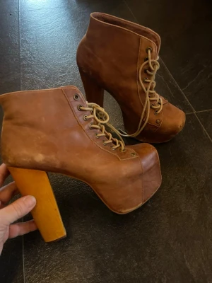 Bruna Campbell platåstövletter med träklack - Säljer ett par bruna stövletter med kraftig platåsula och hög, fyrkantig träklack. Skorna har snörning framtill med metallöljetter och är tillverkade i läder. Perfekta för dig som vill sticka ut med en retroinspirerad look.