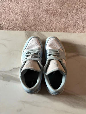Jordan 1 Low vit/blå sneakers - Snygga Jordan 1 Low sneakers i vitt och ljusblått läder med klassisk perforerad tåbox och ikoniska Jumpman-loggan på plösen. Skorna har vita paneler och ljusblå detaljer samt matchande ljusblå snören. Perfekt för dig som gillar streetwear och sportig stil.                                                Pris kan diskuteras privat