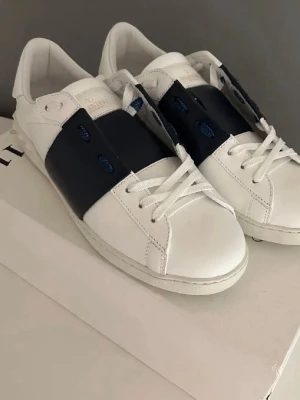 Valentino opens med blå detalj - Snygga vita sneakers från Valentino med bred mörkblå rem över snörningen och coola nitar på hälen. Skorna har en klassisk låg profil och är tillverkade i slätt läder med vita gummisulor. Perfekta för dig som vill ha en stilren men ändå unik look.ÄKTA