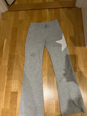 Grå mjukisbyxor med stjärna Kappahl - Grå mjukisbyxor från Kappahl med en stor vit stjärna på ena benet. Byxorna har resår i midjan och är tillverkade i mjukt, bekvämt material. Perfekta för chill dagar hemma eller när du vill ha det extra bekvämt.