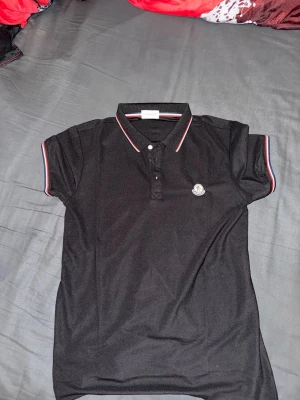 Svart Moncler pikétröja med röd/vit kant - Klassisk svart pikétröja från Moncler med korta ärmar och krage. Tröjan har röda och vita detaljer på krage och ärmslut samt Moncler-logga på bröstet. Perfekt till jeans  för en clean och sportig look.