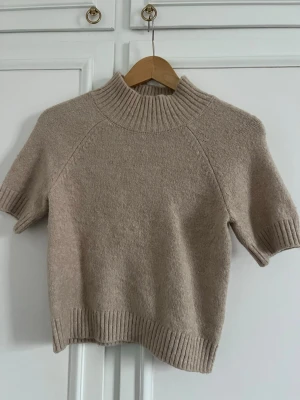 Beige stickad t-shirt med hög krage - Kortärmad stickad tröja i beige med hög ribbad krage och ribbade muddar vid ärmslut och nederkant. Tröjan har en avslappnad passform och är gjord i ett mjukt material, perfekt för lager på lager eller sommarens svalare dagar.