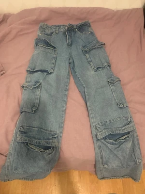 Blå cargobyxor med stora fickor - Säljer ett par blå cargobyxor i denim med flera stora, rejäla fickor längs benen. Byxorna har en lös passform och klassisk cargostil. Perfekta för dig som gillar streetwear och vill ha plats för allt möjligt i fickorna.