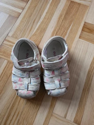 Blommiga sandaler från Gulliver - Söta sandaler från Gulliver i storlek 22. De är vita med blommigt mönster i rosa, lila och blått. Sandalerna har stängd tå, kardborreband och mjuk sula. Perfekta för varma dagar och ger en gullig touch till outfiten.