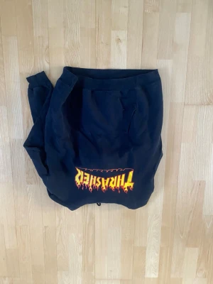 Svart Thrasher hoodie med flames - Säljer en svart hoodie från Thrasher med den klassiska loggan i gult och rött flames-tryck på bröstet. Hoodien har huva och känguruficka framtill. Perfekt för dig som gillar streetwear och skatekultur. Storlek M