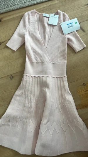 Ljusrosa omlottklänning från Sandro - Supersöt ljusrosa klänning från Sandro med omlott-v-ringning och ribbad struktur upptill. Kjolen har ett subtilt mönster och klänningen har korta ärmar. Perfekt för vår och sommar, i ett mjukt och stretchigt material. Vårens trendigaste  färg!