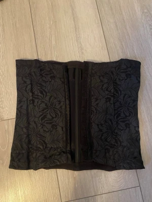 Svart blommig midjekorsett - Säljer en svart korsett med elegant blommönster och hakknäppning framtill. Korsetten har en klassisk form som markerar midjan och är tillverkad i ett tyg med lätt glans. Perfekt för dig som vill ha en snygg siluett och gillar detaljerade mönster.