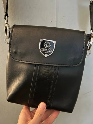 Svart axelväska från Philipp Plein - Snygg svart axelväska från Philipp Plein i slätt konstläder med silverfärgade metalldetaljer och logga framtill. Väskan har justerbar axelrem, lock med magnetknäppning och en praktisk ytterficka med dragkedja på baksidan. Perfekt för att bära det viktigaste. Först till kvarn som gäller säljer den eftersom jag inte har någon användning till den längre👍🏼 pris kan diskuteras vid snabb affär, behöver bara få den såld.