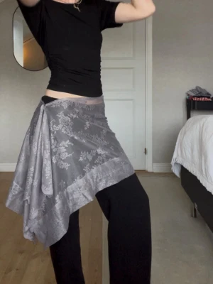 Grå omlottkjol med volang - Säljer en grå handsydd omlottkjol med volang och blommigt mönster. Kjolen har asymmetrisk nederkant och knäpps i midjan för en personlig passform. Perfekt för dig som gillar en unik och flowy stil.  Passar S/M