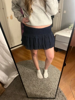 Mörkblå volangkjol med resårmidja - Säljer en mörkblå kortkjol med bred resår i midjan och flera lager volanger. Kjolen har en luftig och flowy känsla, perfekt för dig som gillar en söt och avslappnad stil. Passar till både sneakers och boots. Kan bäras hög och lågmidjad, som kjol eller topp.