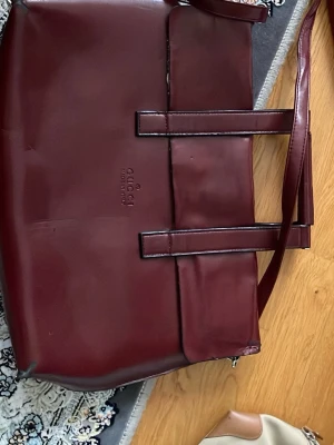 Vinröd handväska från Gucci - Stilren vinröd handväska från Gucci i slätt konstläder. Väskan har dubbla handtag, en stor ytterficka med lock och dragkedja på baksidan. Klassisk rektangulär form och diskret logga framtill. Perfekt för dig som gillar tidlös design och vill ha plats för allt du behöver.den är lite gammal men det går o fixa