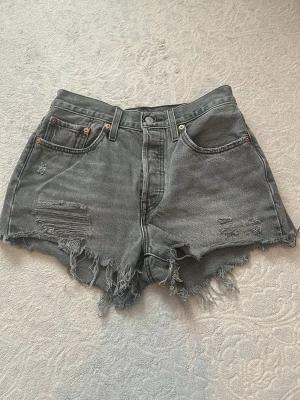 Grå Levi's jeansshorts med fransar - Säljer ett par grå denimshorts från Levi's med hög midja och råa, fransiga benslut. Shortsen har slitna detaljer framtill, klassiska fem fickor och Levi's ikoniska läderpatch bak. Perfekta för sommaren och ger en cool vintagekänsla.