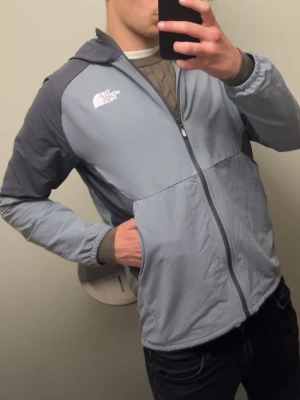  North Face Jacka - Skriv för fler bilder!  Snygg ljusgrå vindjacka från The North Face med mörkgrå kontrastpaneler på axlar och ärmar. Jackan har dragkedja framtill, huva och två sidofickor. Perfekt för blåsiga dagar och enkel att matcha med olika outfits. Jackan är köpt på mos för några veckor sedan så alltså nästan helt ny knappt använd.