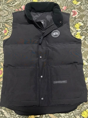 Svart dunväst från Canada Goose - Snygg svart dunväst från Canada Goose med hög krage och två stora framfickor. Västen har ett broderat Canada Goose Arctic Program-märke på bröstet och stängs med både dragkedja och tryckknappar. Perfekt för lager-på-lager och streetwear. PRISET KAN DISKUTERAS!!!!!