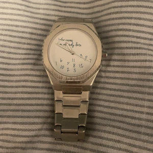 Silvrig klocka med textad urtavla - Stilren klocka i silverfärgad metall med länkarmband. Urtavlan är vit med handskrivna siffror och texten 'Who cares I'm already late' vilket ger en lekfull touch. Perfekt accessoar för dig som gillar unika detaljer. Extra länkar och låda ingår! Passa på och köp fler plagg/accessoarer så får du en bundle rabatt!