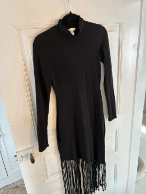 Svart långärmad klänning H&M - Svart långärmad klänning från H&M med hög krage och fransdetaljer längst ner. Klänningen har en smal passform och är tillverkad i mjukt material som känns bekvämt mot huden. Perfekt för dig som gillar en stilren look med en twist.