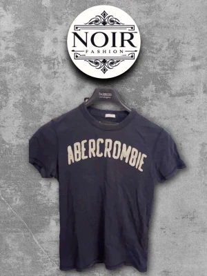 Mörkblå Abercrombie t-shirt - Snygg mörkblå t-shirt från Abercrombie med vit text framtill. Klassisk rund halsringning och korta ärmar. Perfekt till jeans eller shorts för en avslappnad look.