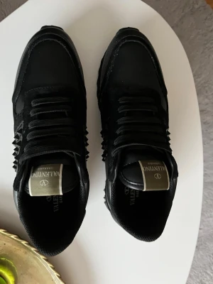 Svarta Valentino Garavani sneakers - Snygga svarta sneakers från Valentino Garavani med coola nitar på sidorna. Skorna har en mix av mocka och läder, samt svarta skosnören och en robust sula. Perfekta för dig som gillar en edgy och exklusiv stil. Använda 1 gång👍 Kan dra ner priset🔥
