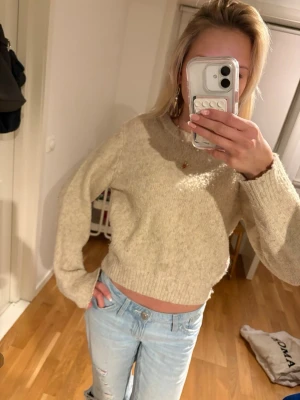 Beige stickad cropped tröja - En beige stickad tröja med rund hals och långa ärmar. Tröjan har en avslappnad passform och ribbstickade muddar vid ärmslut och nederkant. Perfekt att matcha med jeans för en cool look.