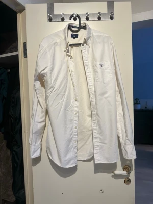 Vit skjorta från GANT med ficka - Klassisk vit skjorta från GANT med bröstficka och broderad logga ovanför fickan. Skjortan har lång ärm, knappar framtill och en tidlös design som passar till det mesta. Tillverkad i mjuk bomull för en bekväm känsla.