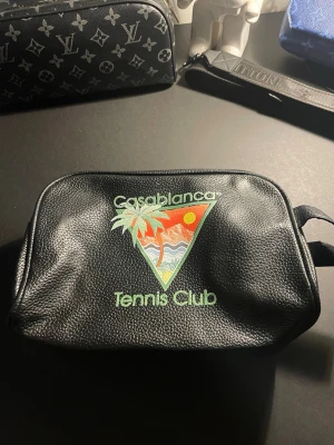 Casablanca Tennis Club svart necessär - Säljer en svart necessär från Casablanca med tryck av en färgglad triangel med palm, sol och vågor samt texten 'Casablanca Tennis Club' på framsidan. Tillverkad i strukturerat konstläder och har en enkel, rektangulär form med dragkedja.