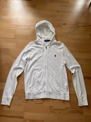 Vit hoodie från Polo Ralph Lauren - Vit hoodie med dragkedja från Polo Ralph Lauren. Klassisk design med två fickor fram och broderad logotyp på bröstet. Mjuk bomullskänsla och huva med snören. Perfekt för en chill och stilren look.