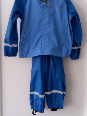 Barn - Blått regnställ från Exani - Storlek 104 - Snyggt blått regnställ från Exani med reflexdetaljer på ärmar och ben. Setet består av jacka och byxor i vattenavvisande material, perfekt för blöta dagar. Jackan har dragkedja och tryckknappar, samt praktiska fickor. Byxorna har resår i benslut och fotstrappar. Storlek 104 är en standardiserad barnstorlek (centilong) som motsvarar en kroppslängd på 104 cm, vilket oftast passar barn i åldern 3–4 år.