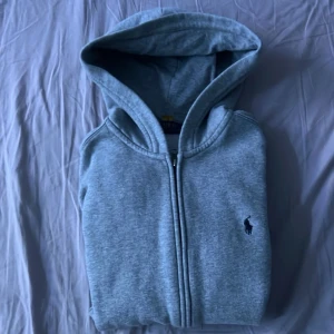 Grå hoodie från Polo Ralph Lauren - Snygg grå hoodie från Polo Ralph Lauren med dragkedja framtill och klassisk huva. Ikoniska lilla Polo-loggan broderad på bröstet. Tillverkad i mjukt och bekvämt material, perfekt för en chill och avslappnad stil.