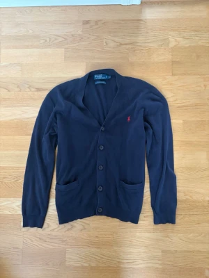 Mörkblå cardigan från Polo Ralph Lauren - Klassisk mörkblå cardigan med knappar från Polo Ralph Lauren. Tröjan har två fickor framtill och den ikoniska röda loggan broderad på bröstet. Tillverkad i mjukt material och har lång ärm och ribbade muddar. Storlek L men sitter som M. Pris går att diskuteras✅. 
