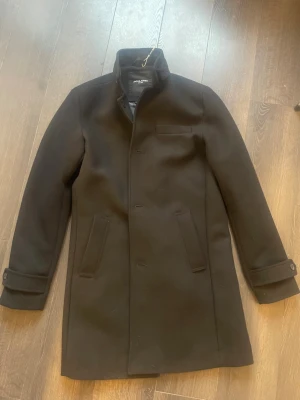 Svart klassisk ullkappa Jack & Jones - Stilren svart kappa från Jack & Jones i ullblandning. Rak passform med dold knäppning framtill, två snedställda fickor och en bröstficka. Kappan har hög krage och dekorativa spännen vid ärmsluten. Perfekt för en snygg och tidlös look. Använd max 10 gånger!