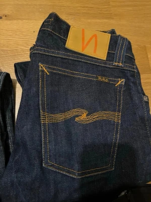 Mörkblå jeans från Nudie Jeans - 399 styck Säljer två par klassiska mörkblå jeans från Nudie Jeans med ikonisk orange söm på bakfickan och läderpatch i midjan. Jeansen har rak passform och är tillverkade i slitstarkt bomullsdenim. Perfekta för dig som gillar stilrena och tidlösa jeans.