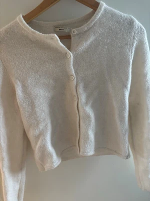 Vit kofta från Ginatricot med knappar - Mysig vit kofta från ginatricot med rund halsringning och fyra knappar framtill. Koftan är stickad i ett mjukt material och har lång ärm samt en något kortare, boxig passform. Perfekt att slänga över en topp för en enkel och stilren look.