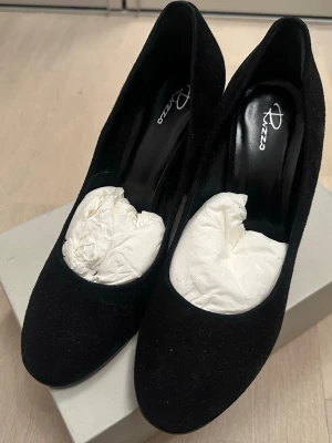 Rizzo Pumps i svart mocka, storlek 39 - Stilrena svarta pumps från Rizzo i mjuk mocka med rundad tå och bred klack. Klassisk och tidlös design som passar till många olika looks. Skorna har en bekväm innersula och en stadig klack som ger bra stöd.
