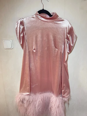 Rosa sammetsklänning med fuskpäls - Unik rosa kortklänning i glansigt sammetstyg med höga axelvaddar och mjuk fuskpälsdetalj längst ner. Klänningen har en hög krage och puffiga axlar som ger en cool retrovibe. Perfekt för dig som vill sticka ut!  Fint skick, inga skavanker. Weekday, storlek S. Djur, rök och parfymfritt hem.