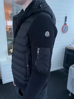 Monclear jacka  - Svart cardiganjacka från Moncler med vadderad front och stickade ärmar. Jackan har huva, dragkedja framtill och en ficka med dragkedja på ärmen. Moncler-logga på vänster ärm och ribbade muddar. Perfekt för en trendig lager-på-lager-look. Nypris 12800kr. Tags finns vid inköp i butik. 