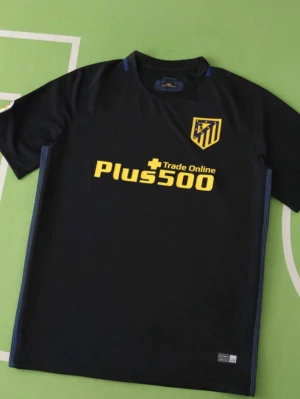 Atlético Madrid | Svart matchtröja - Snygg svart matchtröja från Atlético Madrid med gula detaljer och klubbmärke på bröstet. Trycket 'Plus500 Trade Online' i gult framtill. Tillverkad i ett lätt och ventilerande material, perfekt för fotboll eller träning.