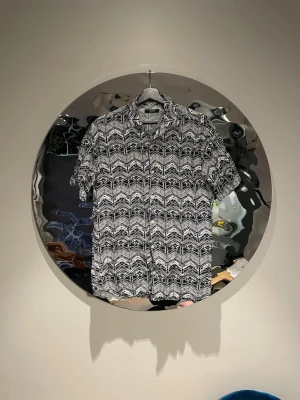 Svartvit mönstrad skjorta med knappar - Säljer en svartvit skjorta med korta ärmar och ett grafiskt, zigzag-rutigt mönster. Skjortan har klassisk krage och knappar framtill. Perfekt för dig som gillar att sticka ut med coola mönster och enkel stil.