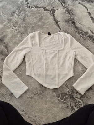 Vit långärmad topp från SHEIN - Trendig vit långärmad topp från SHEIN med fyrkantig urringning och markerade sömmar framtill. Modellen är croppad och har en figurnära passform som framhäver midjan. Perfekt att styla med höga jeans eller kjol.